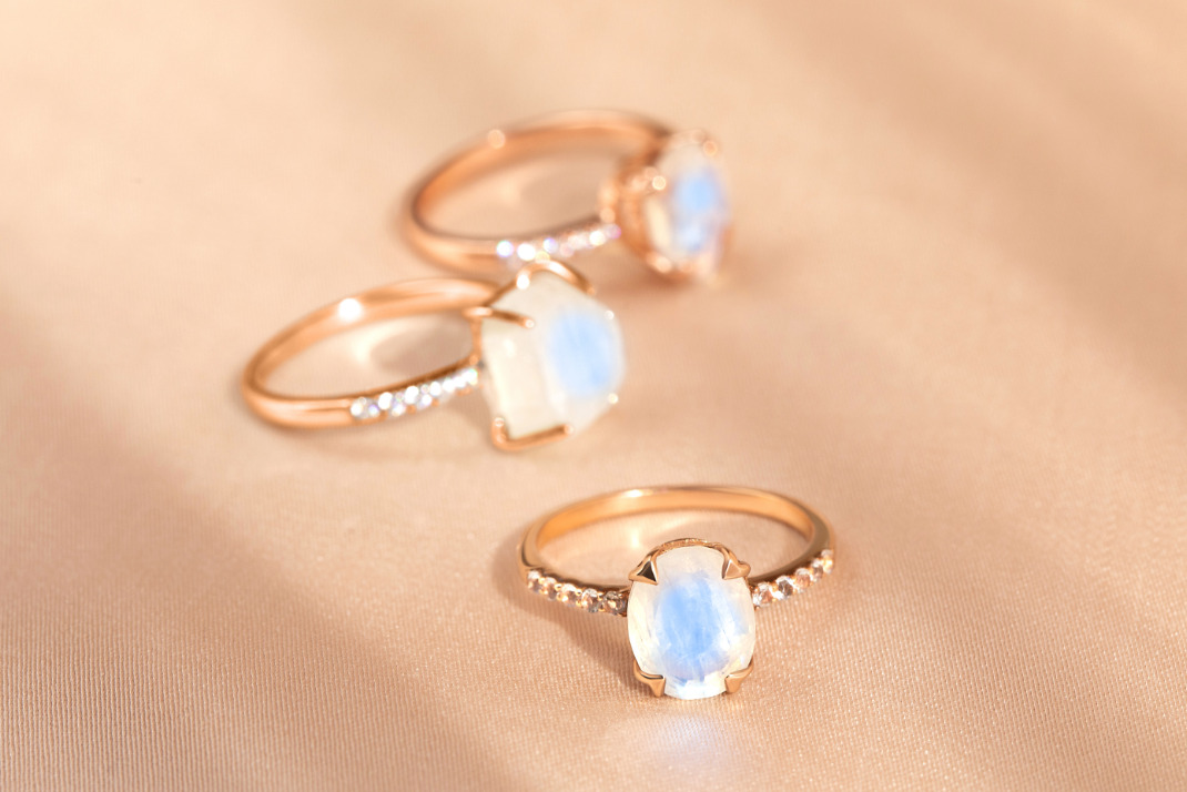 Three rose gold rings with Moonstone gems are presented on a beige surface.