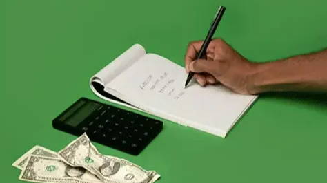 Green surface with a white notebook, black calculator, and three $1 USD bills. A hand is holding a pen and writing notes in the white notebook.