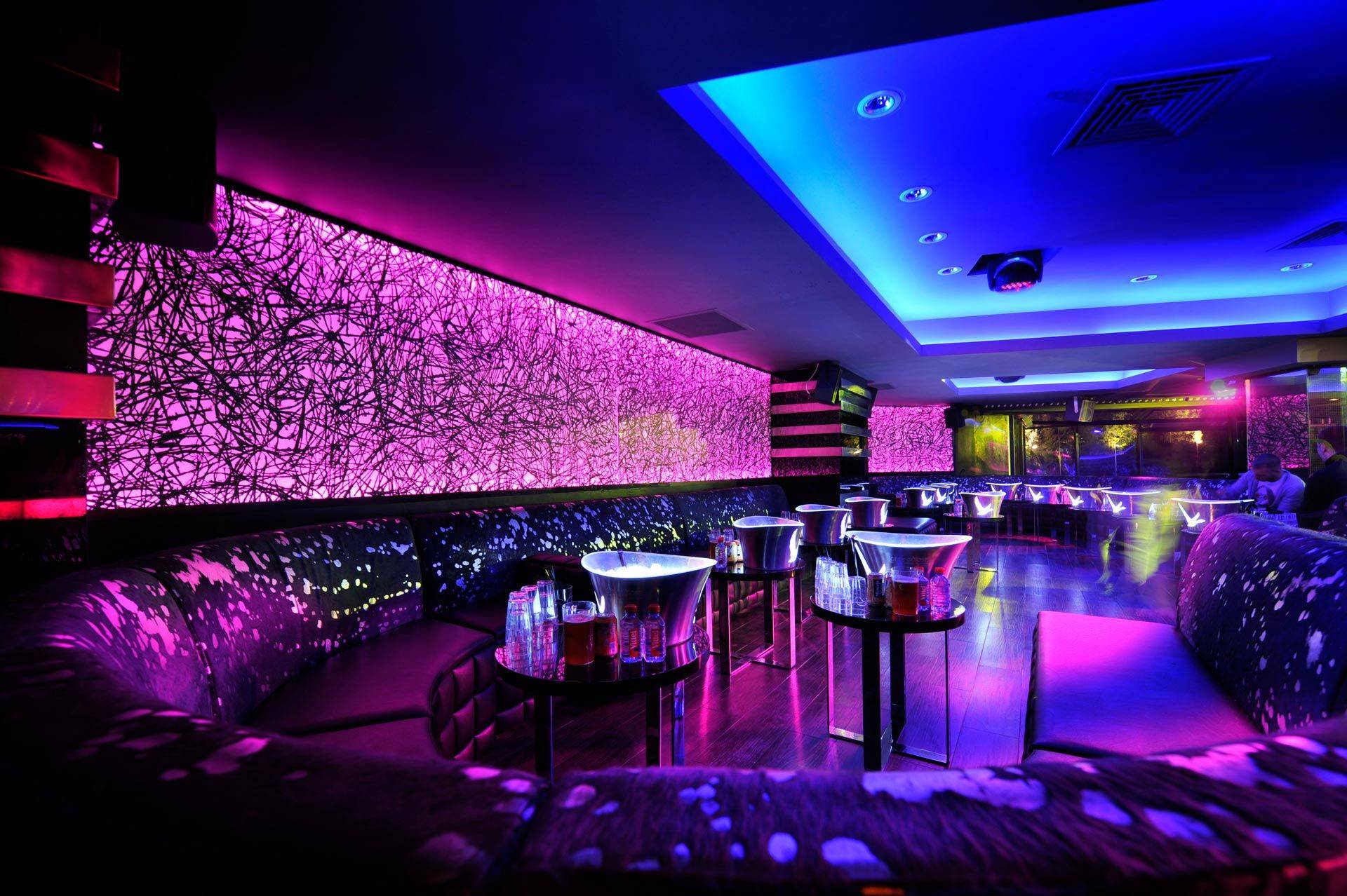 Vibrant nightclub interior with purple and blue lighting, featuring abstract wall patterns, modern curved seating, and illuminated tables set with bottles and ice buckets.