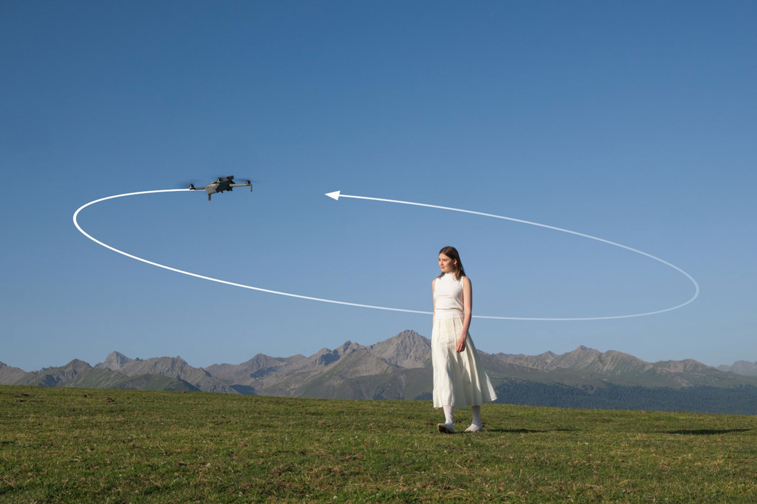 A woman in white stands on grass with a DJI Mini 5 Pro drone flying nearby.