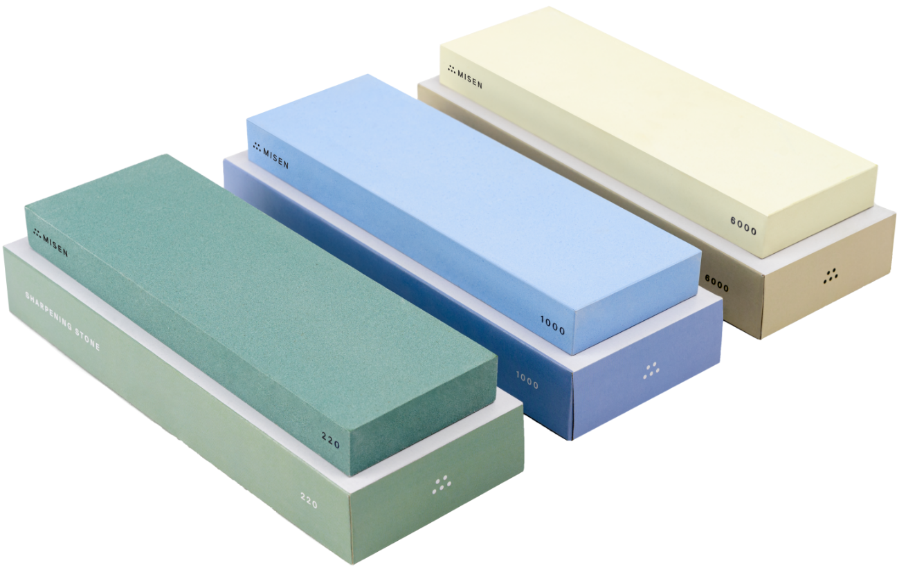 A green Misen 220-grit sharpening stone, a blue Misen 1000-grit sharpening stone, and a cream Misen 6000-grit sharpening stone laid side by side atop their storage boxes.