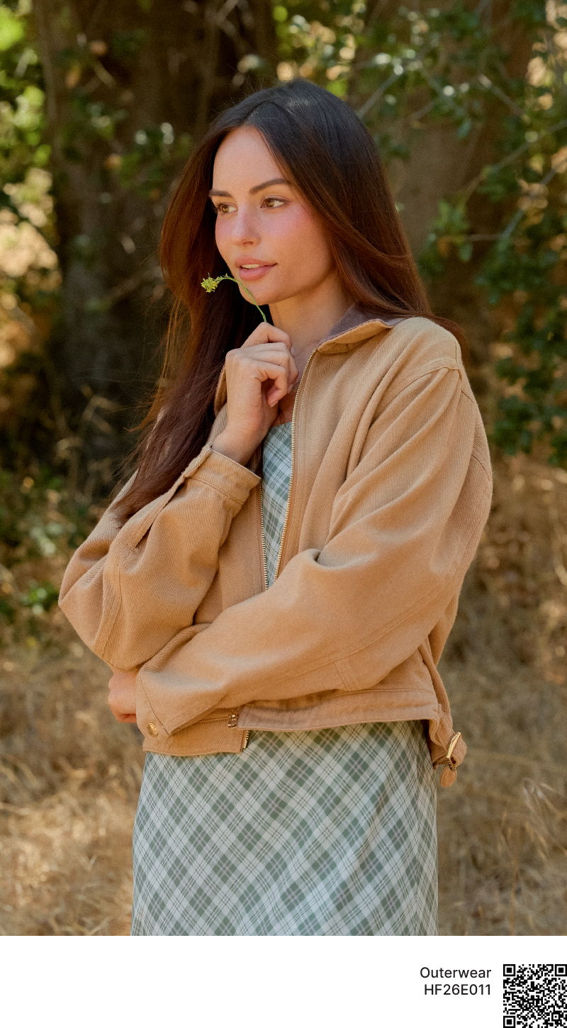 Young woman in a brown jacket standing outdoors, smiling with a natural background.