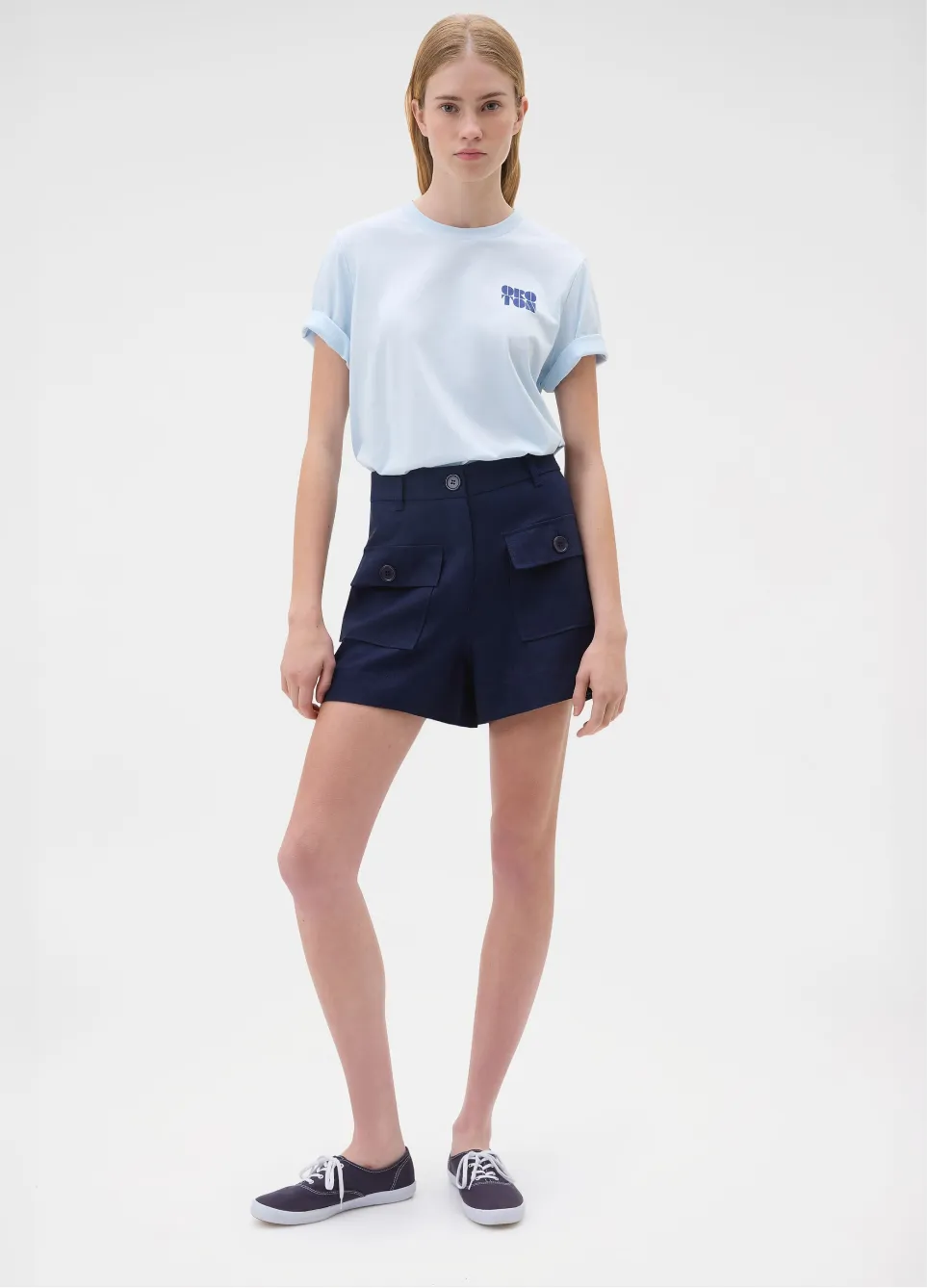 Young woman in light blue shirt and navy shorts standing confidently.