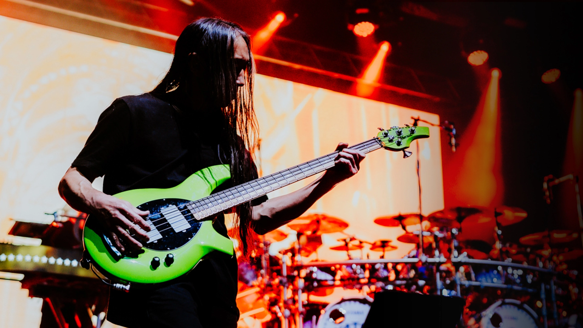 John Myung playing a green Music Man Bongo bass guitar on stage with drums in background.