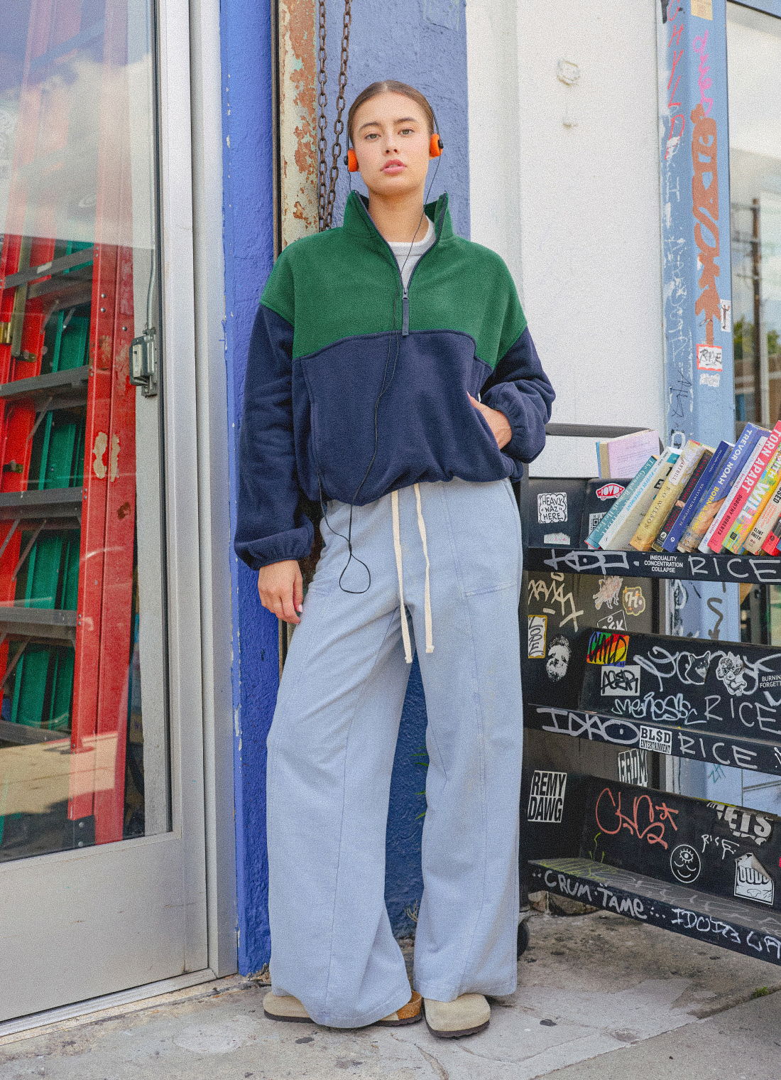 Person wearing a green and navy pullover with light blue pants, standing outside a store.
