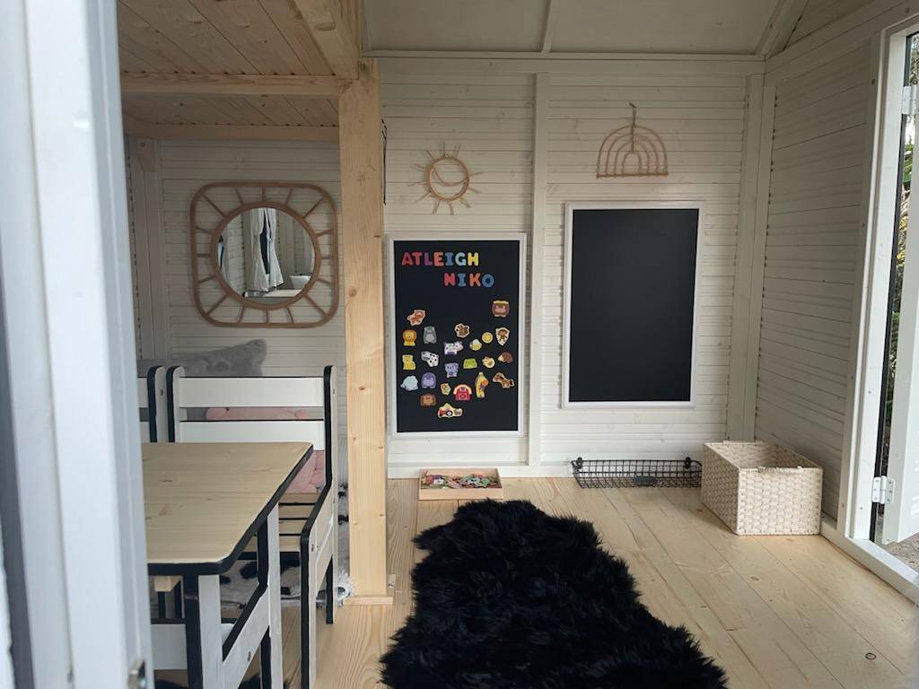 Kids White and Black Outdoor Playhouse inside view with kids furniture, mirror, blackboard and black rug on a wooden floor by WholeWoodPlayhouses