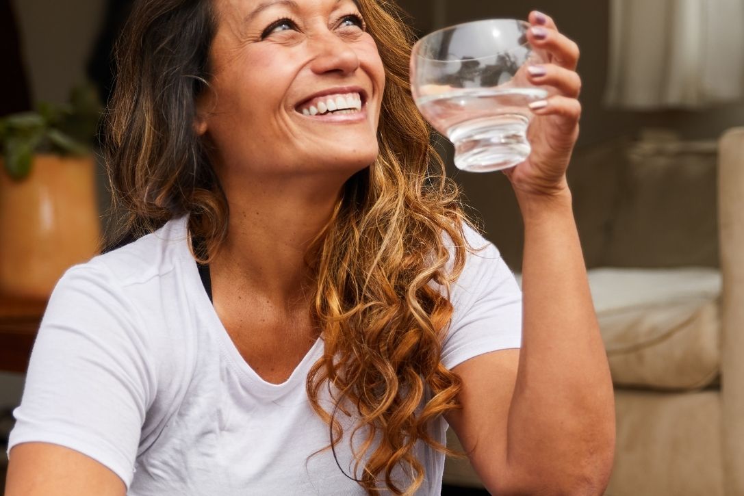 Smiling woman holding a glass of water, sitting in a cozy environment.