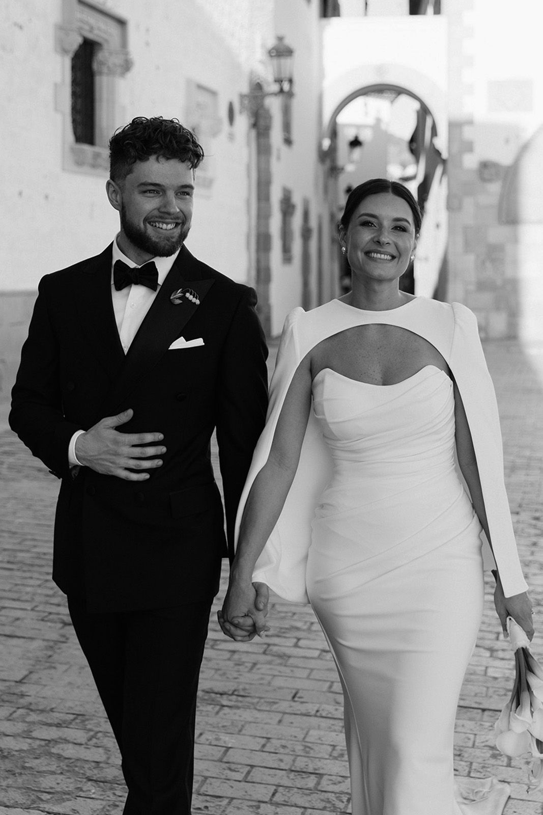 Couple walking hand in hand in elegant attire on a cobblestone street.