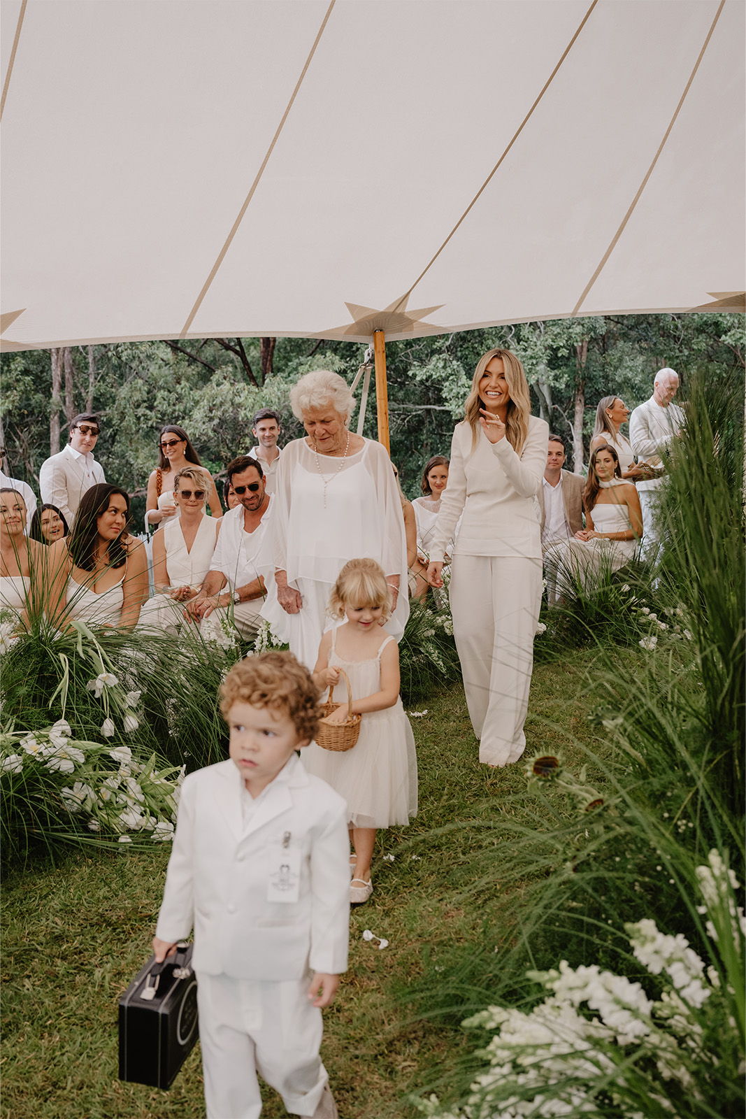 Children and adults in white attire walking down an outdoor aisle at a wedding.
