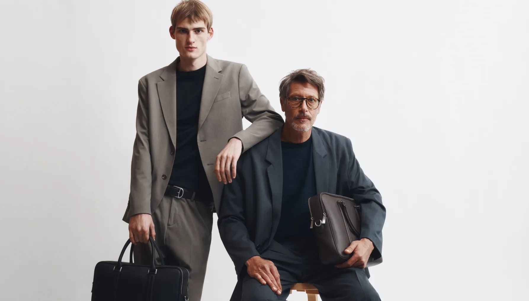 Two men in suits posing, one standing and one sitting, holding briefcases.