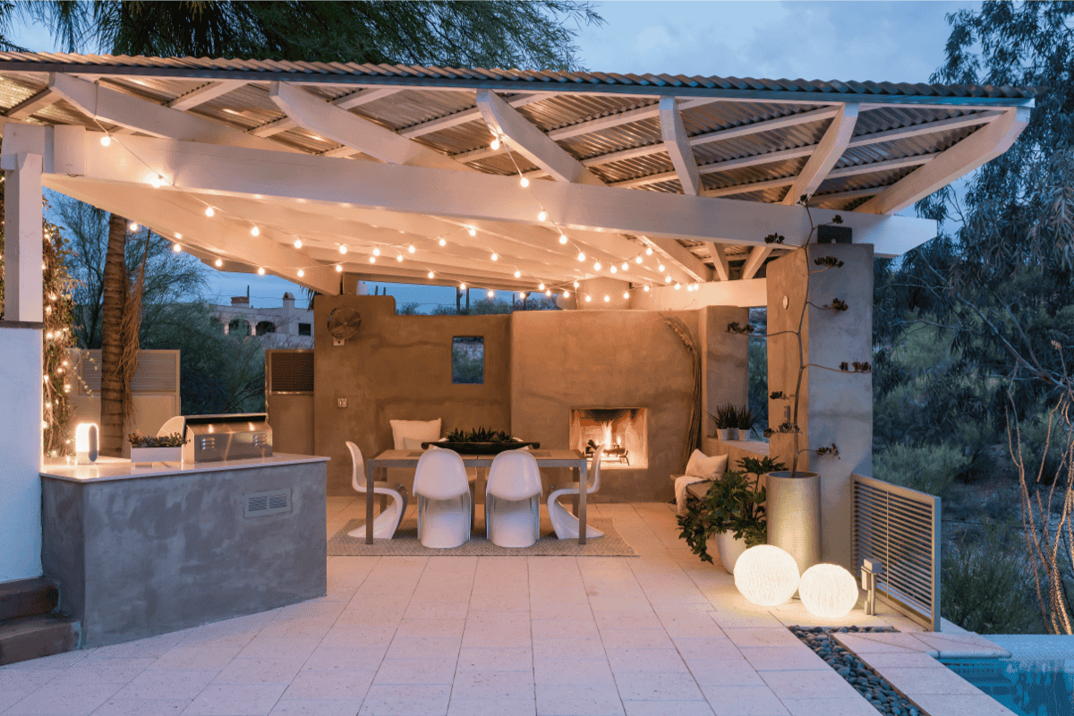 Covered outdoor kitchen and dining pavilion with string lights and fireplace, designed for evening use in the desert.