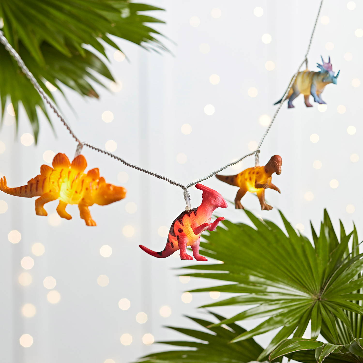 Dinosaur fairy lights in a white room with curtain lights behind them and plants around them.