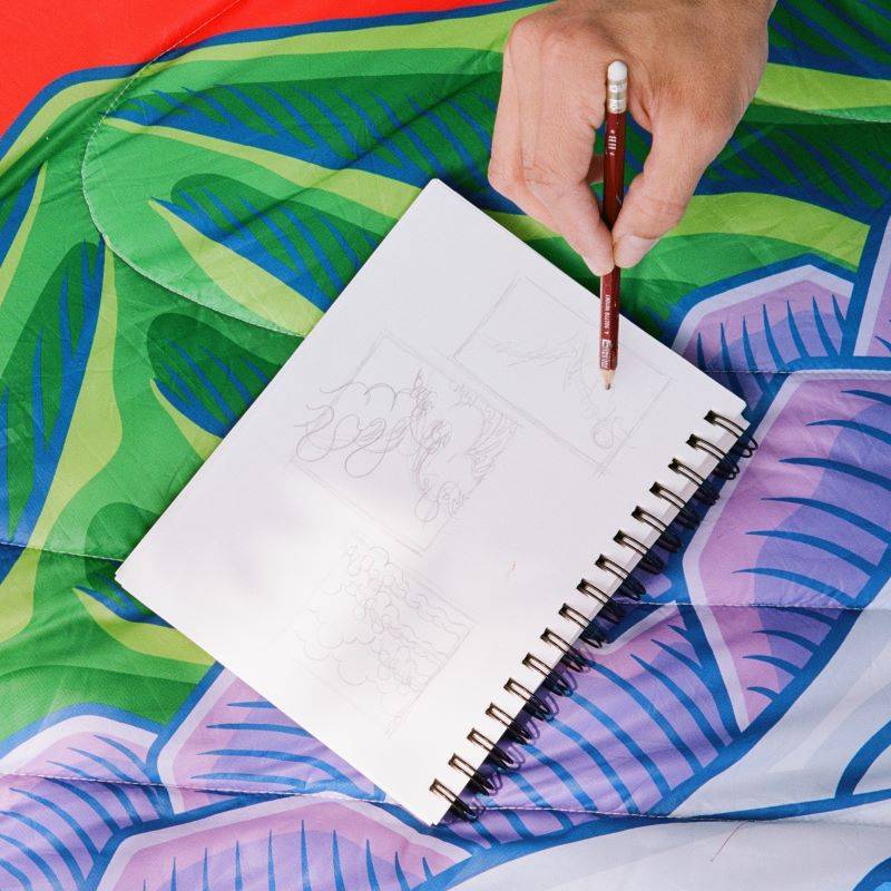 A close up of Aaron Kai's hand drawing some some rough  sketches on top of the Rumpl Original Puffy Blanket with his print, the Hokusai Great Wave. 