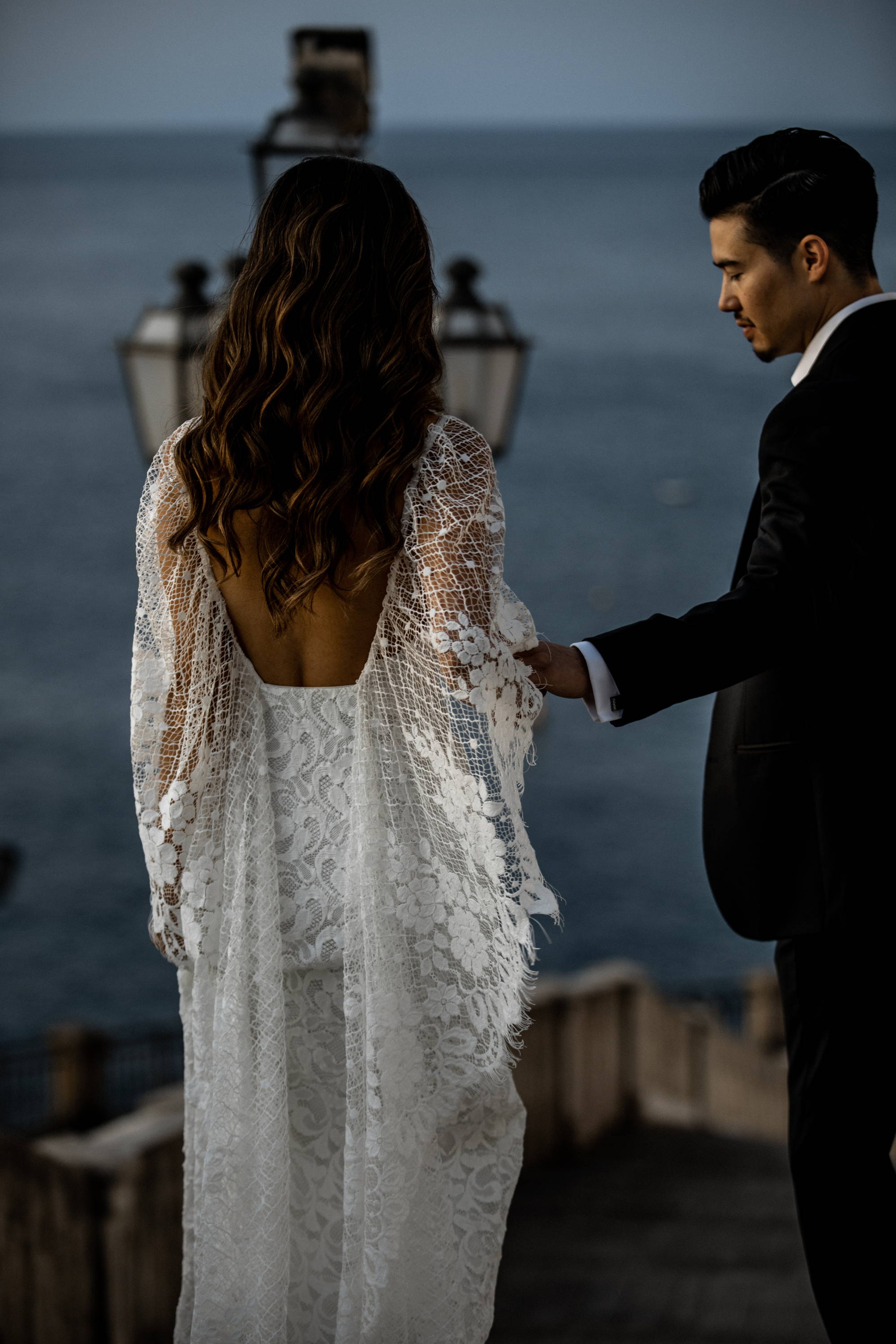 La mariée portant une robe en dentelle drapée sur l'épaule et le marié portant un costume noir.