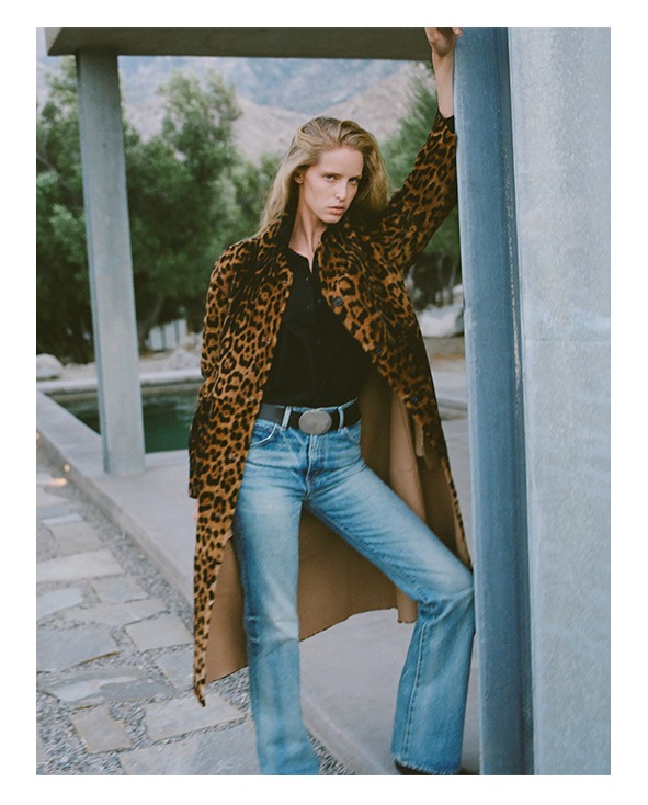 Woman in leopard coat and flared jeans, posing outdoors by a pool.