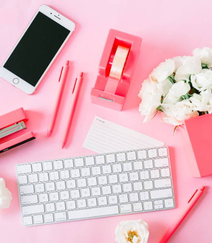 Neon coral desk set with a tape dispenser, stapler, pens, keyboard, and iphone on a pink table.