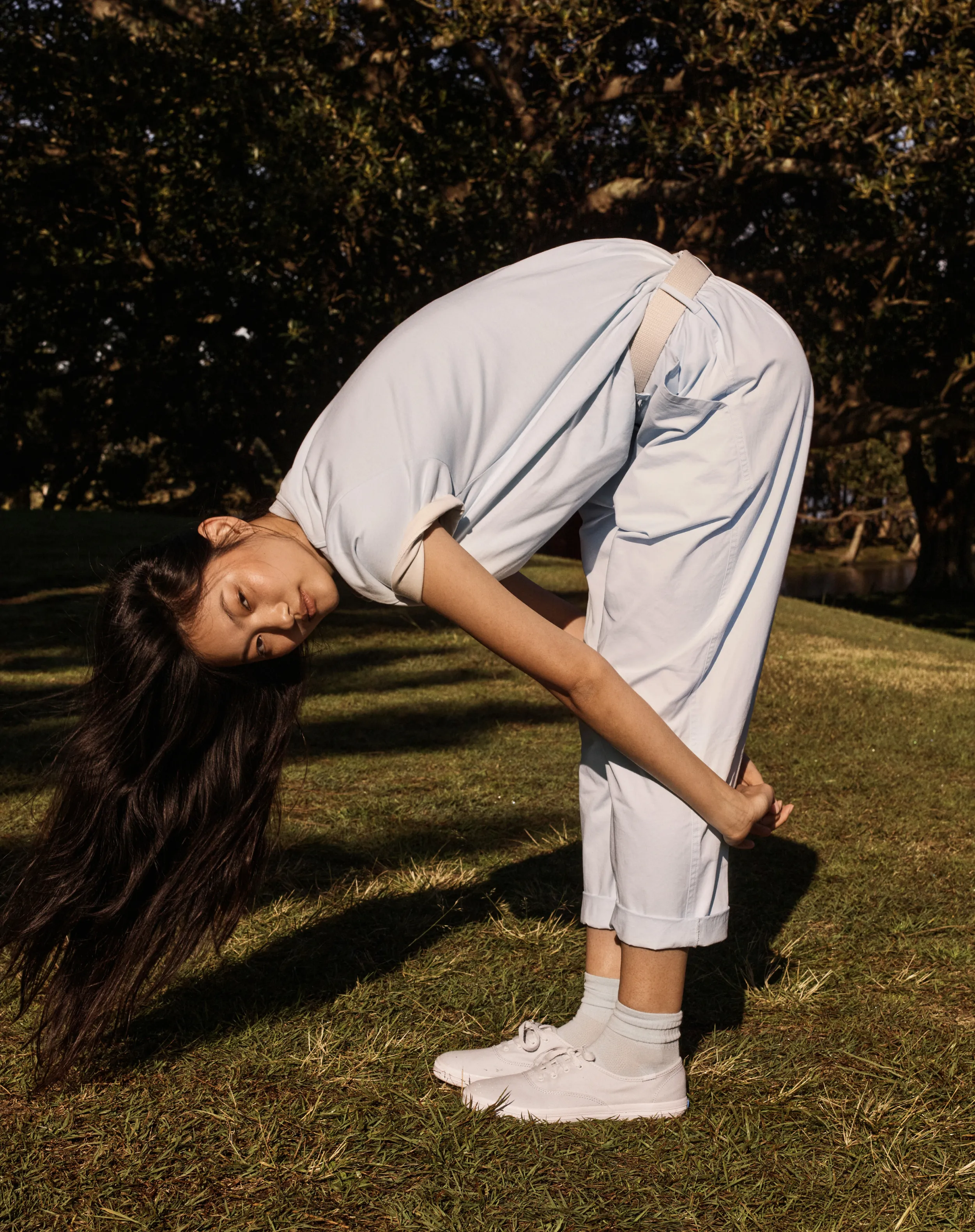 Person bending forward in a grassy area, wearing light clothing and shoes.