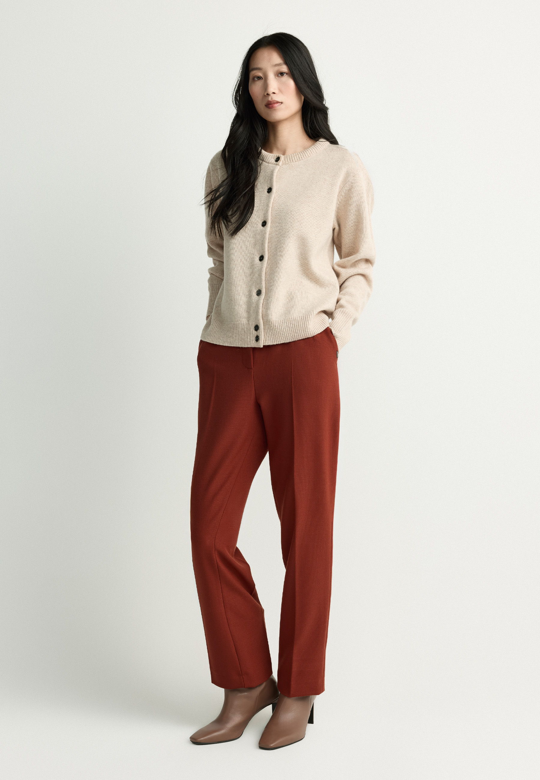 A woman in a beige cardigan and red pants stands against a light background.