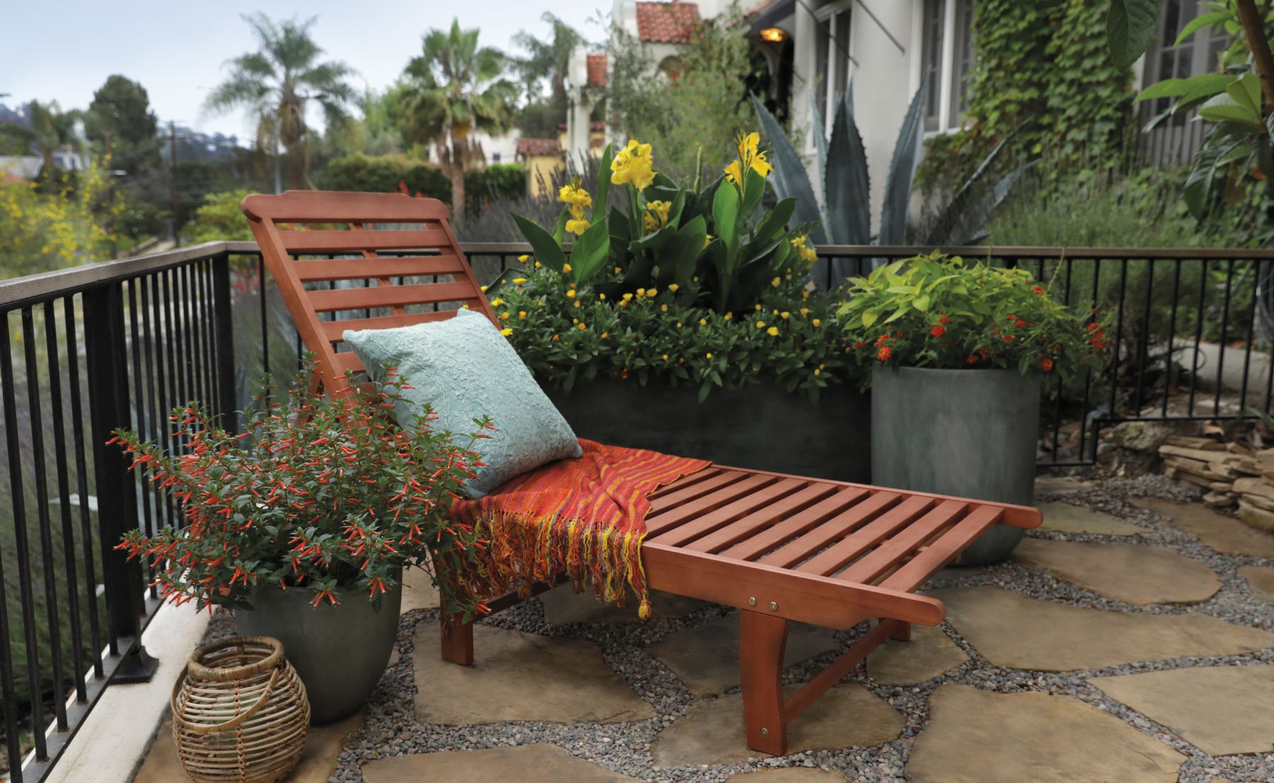 Patio container garden featuring Vermillionaire® Large Firecracker Plant with vibrant orange-red tubular blooms, yellow canna flowers, and chartreuse foliage surrounding a wooden lounge chair.