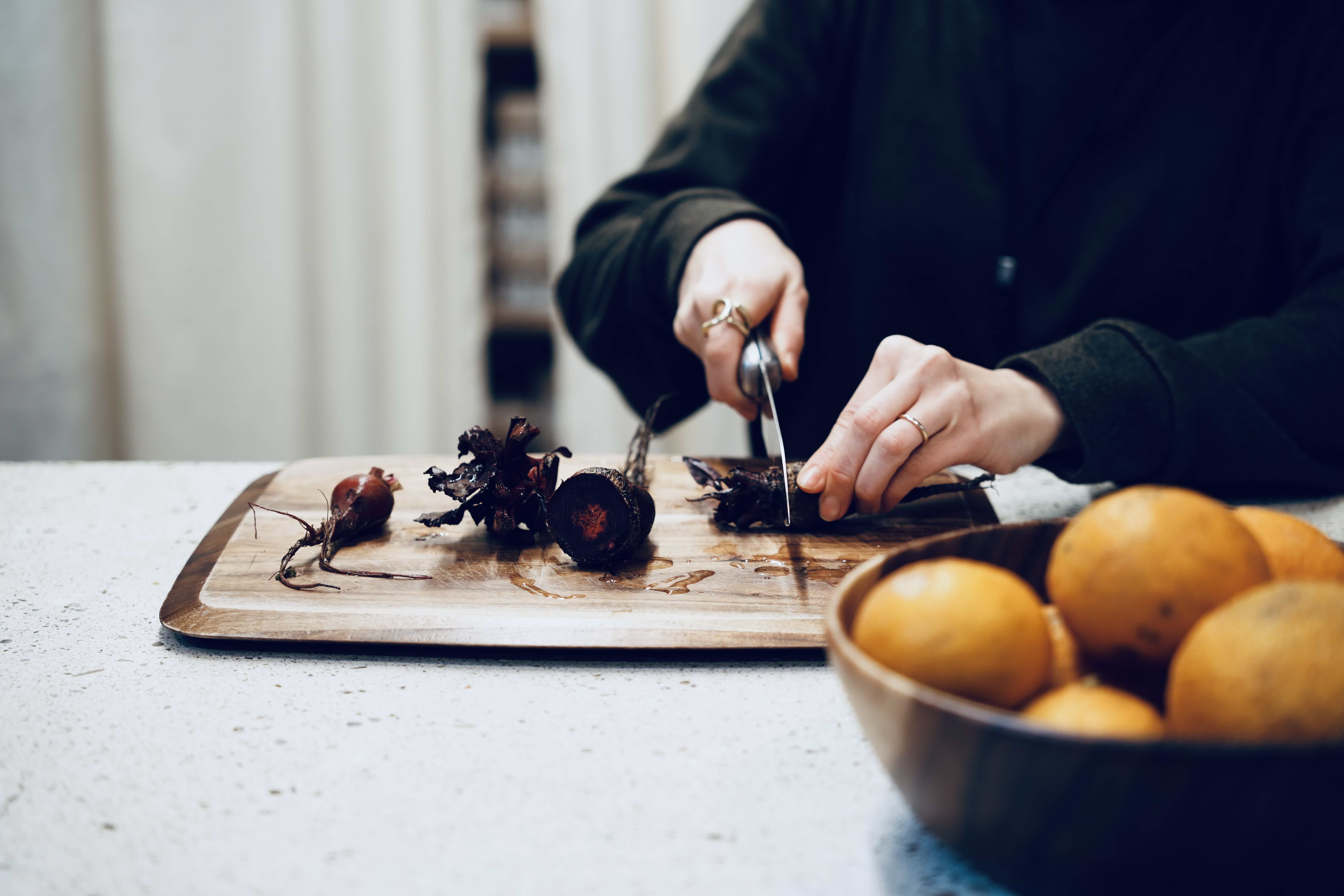 Person slicing food on a wooden cutting board with oranges in a bowl.