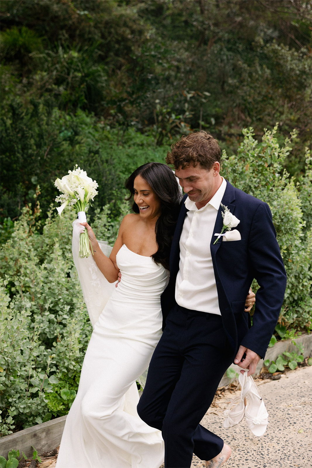 A couple joyfully walking together, holding flowers, in a lush outdoor setting.