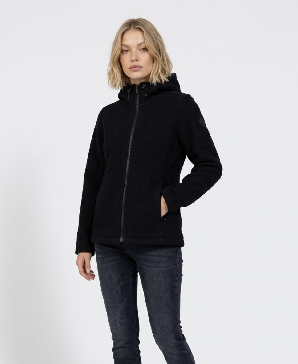 Woman wearing a black zip-up jacket and jeans, standing against a plain background.