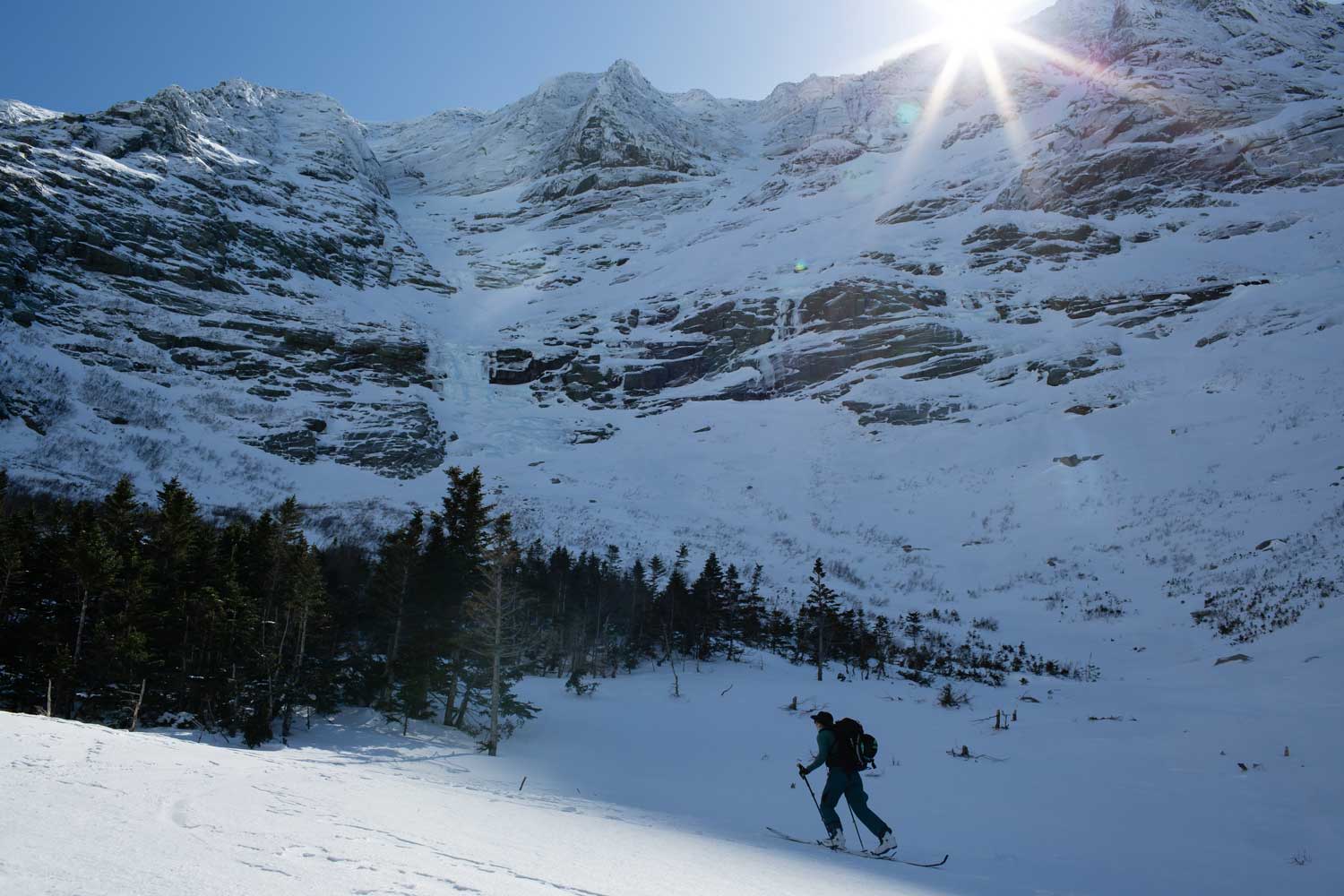 Person splitboarding snow-covered terrain with mountains and sun in background.