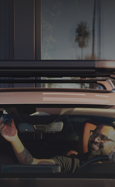 Person inside a car, holding a camera, smiling at the viewer.