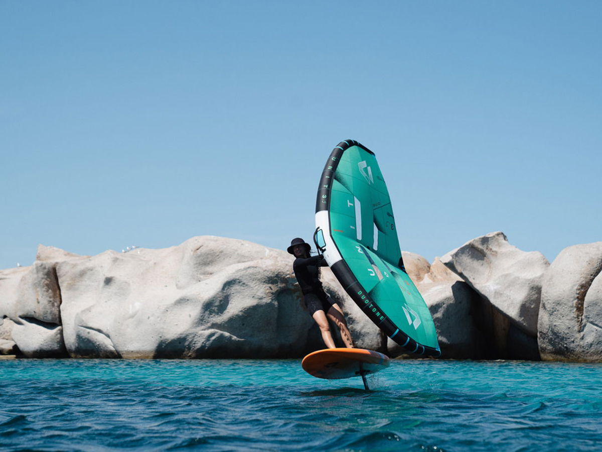 Person foil surfing on water near large rock formations under a clear blue sky.