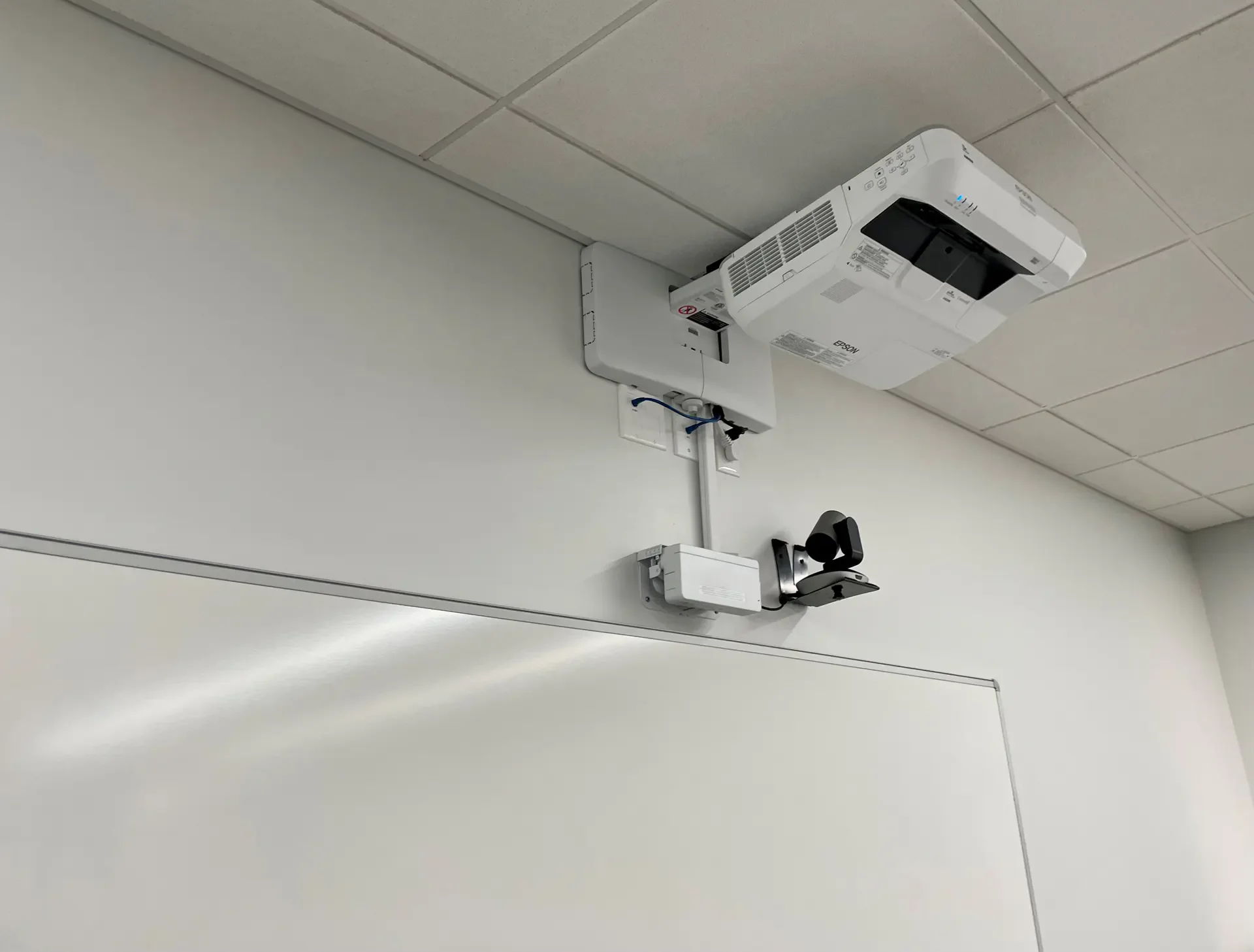 Projector and camera mounted on a white wall above a blank whiteboard.