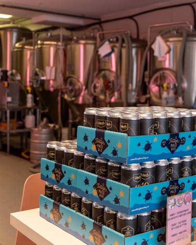 Stack of canned beverages in a brewery setting with brewing equipment in background.
