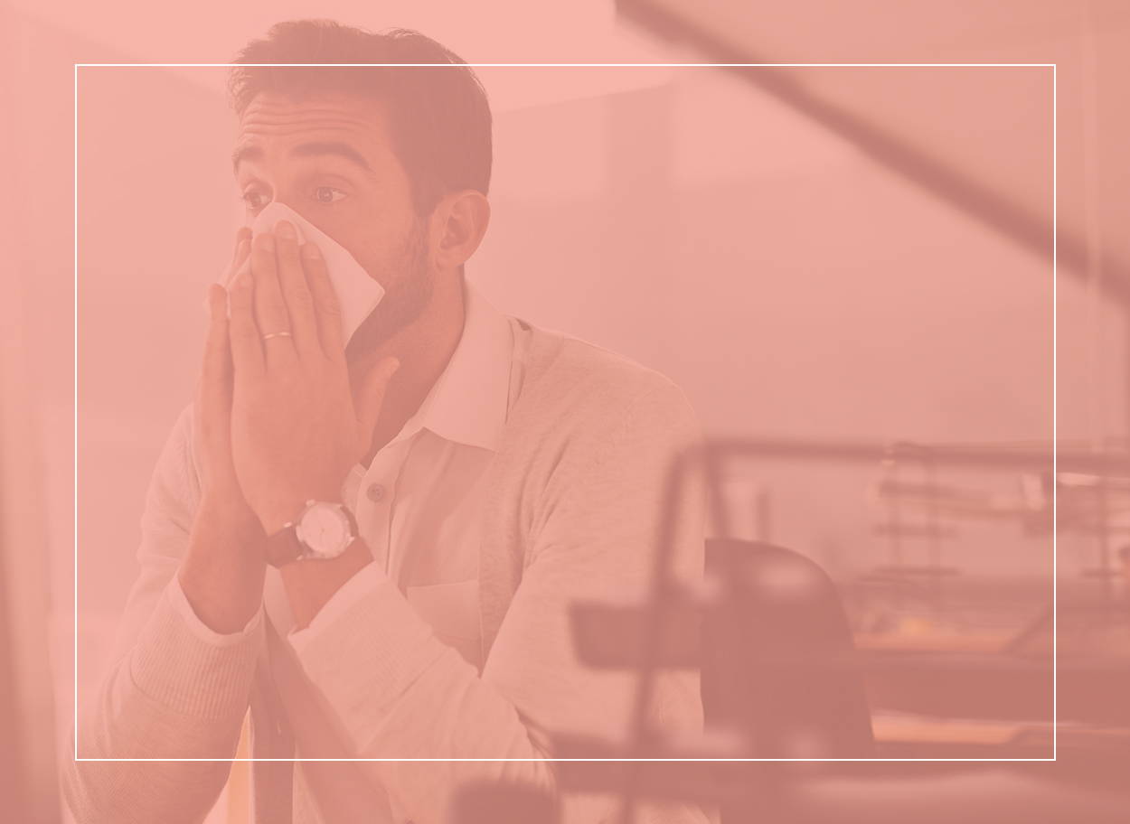 Homme en tenue de travail se mouchant à son bureau. Les symptômes d’une allergie aux acariens font qu’il lui est difficile de se concentrer sur son travail.
