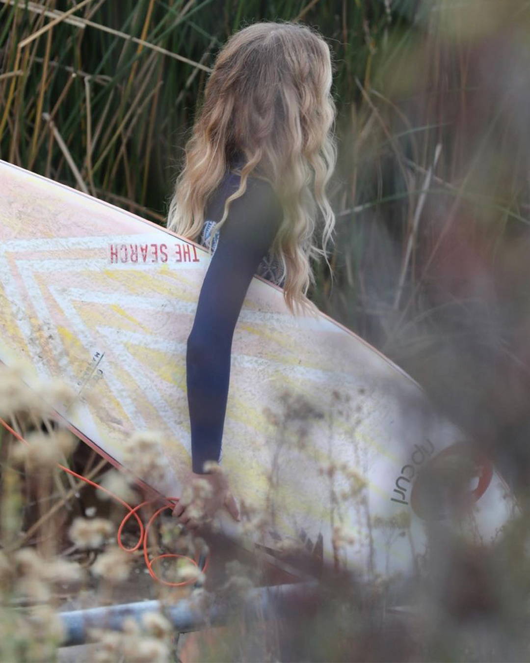  Image of a surfer holding a surfboard taken through greenery.