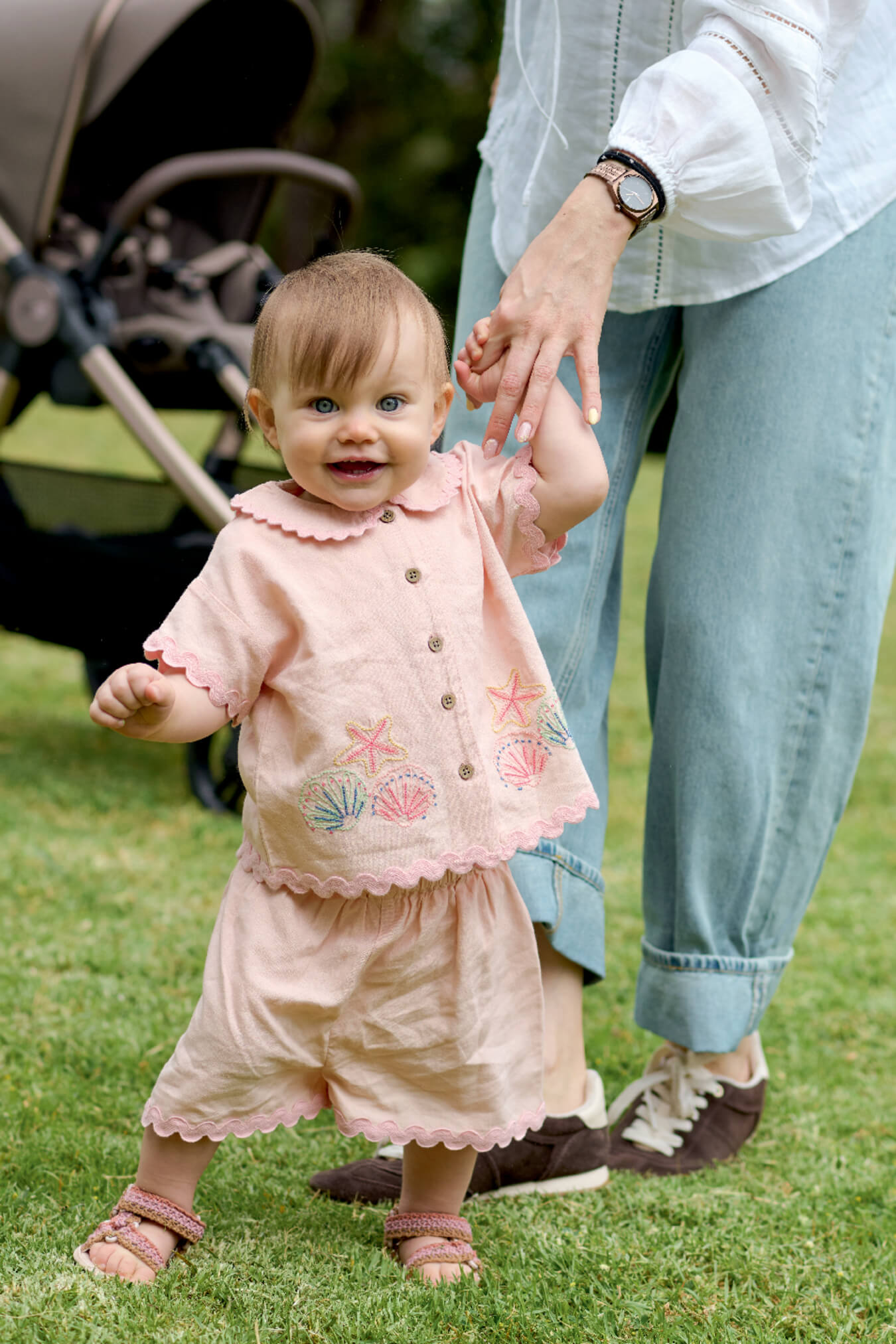 A toddler in a pink outfit holding an adult's hand on grass.
