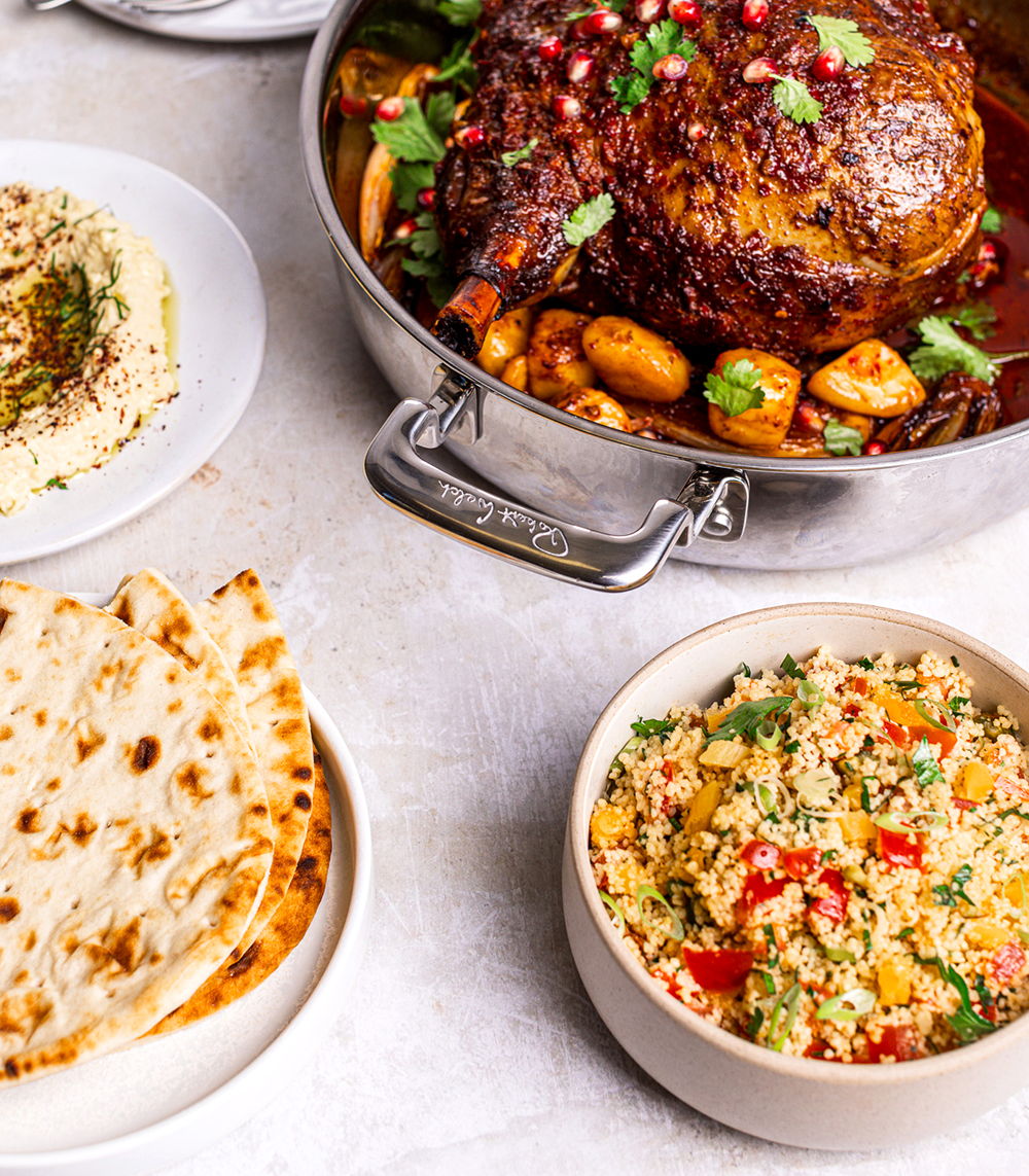 A feast with roast lamb, couscous, hummus, and flatbread on a table.