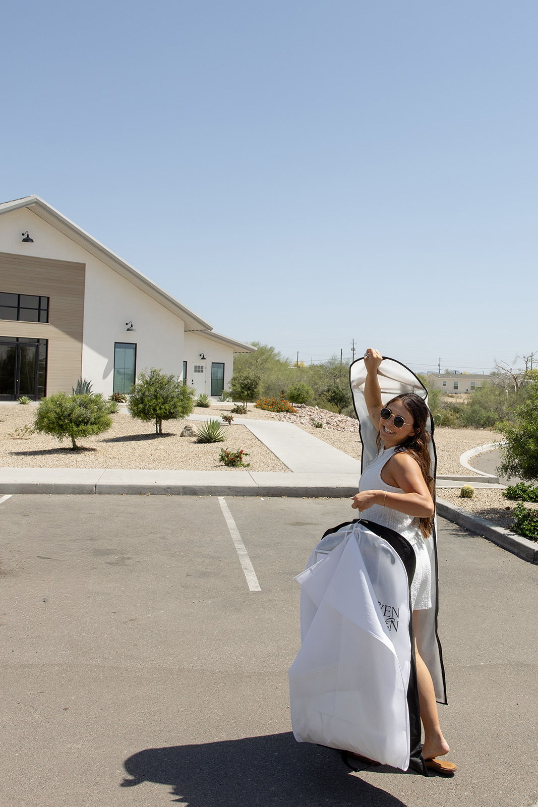 Woman in a white dress poses outside a modern building on a sunny day.