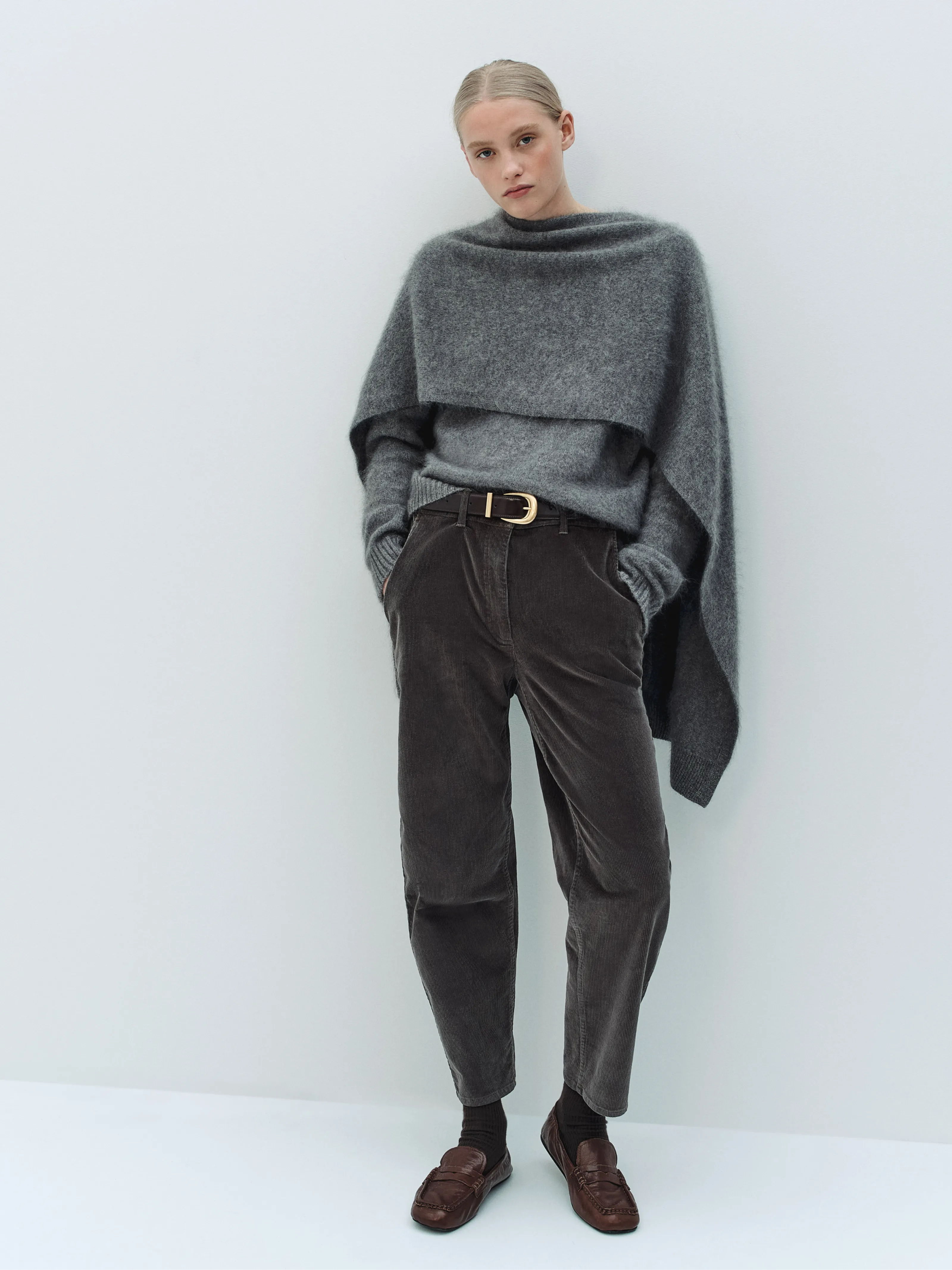 Woman in a gray sweater and brown pants, standing against a light background.