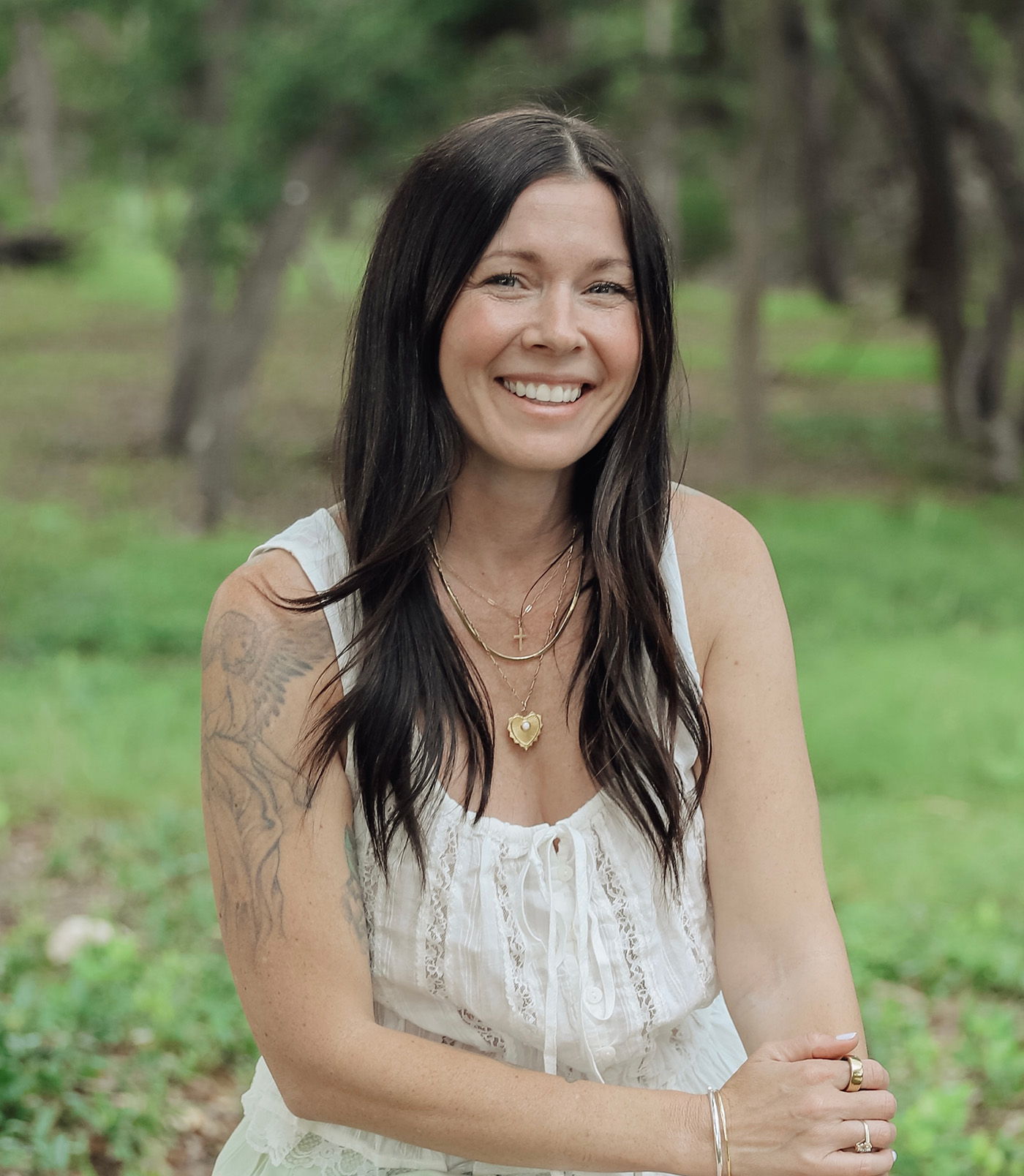 Smiling woman with long hair sitting outdoors in a green setting.