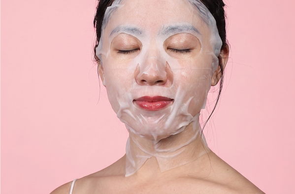 Woman with a facial mask on her face, eyes closed, against a pink background.