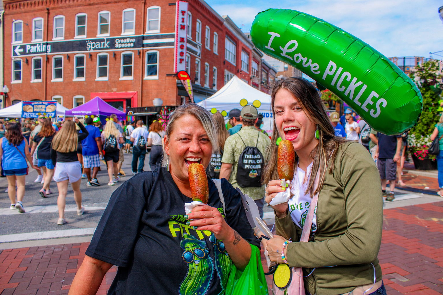 Two women holding food, smiling with a pickle-themed balloon at a festival.