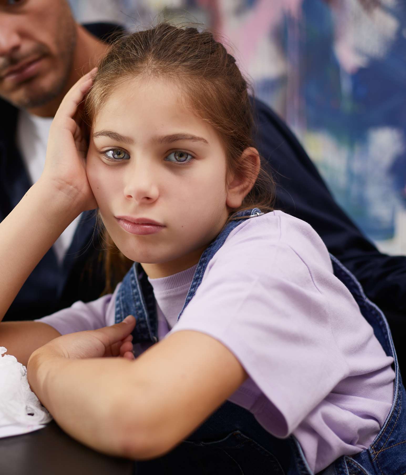 Girl leaning sadly on her elbow, doing homework with her dad – she has allergies and there are crumpled tissues on the table 