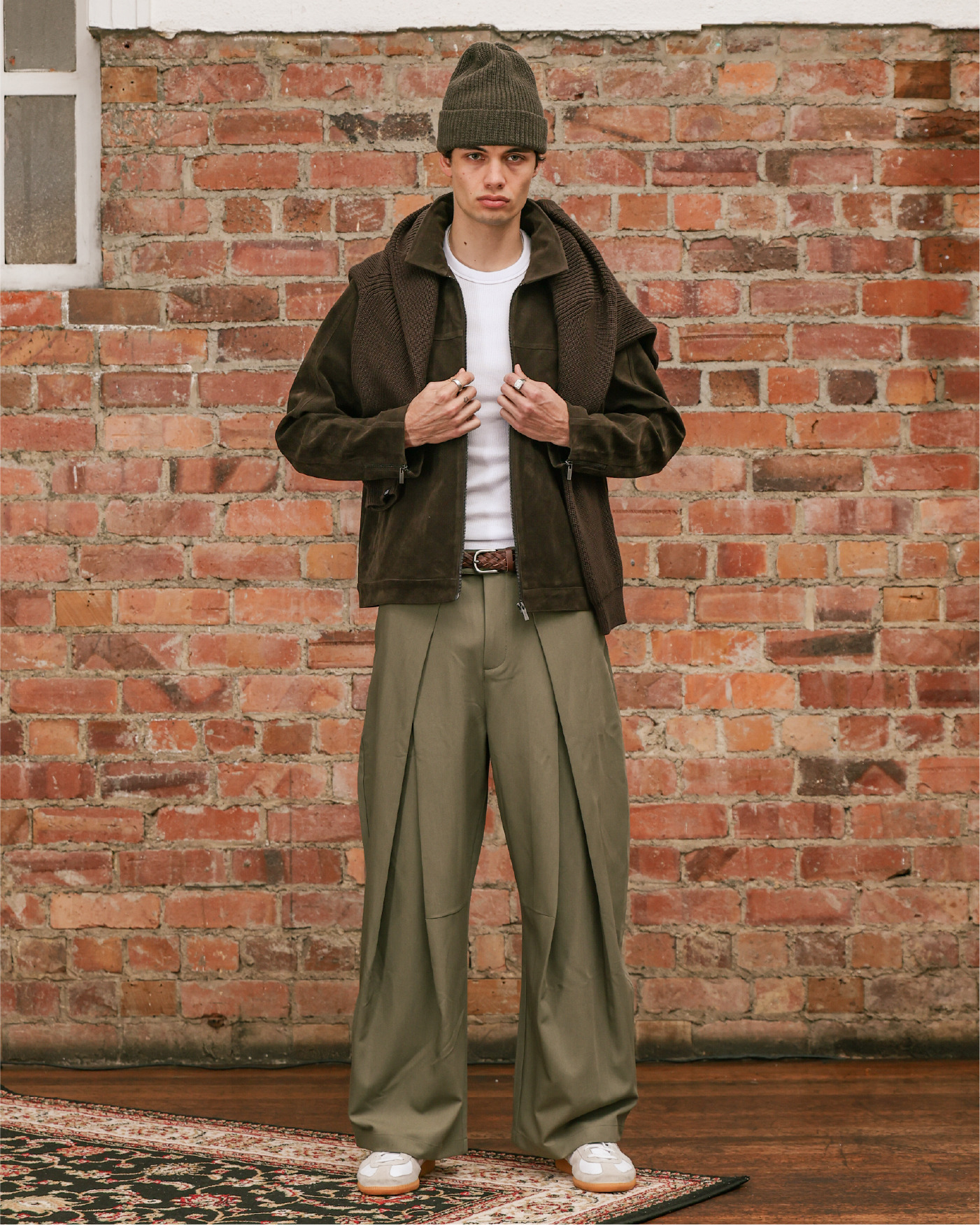 Person wearing a brown jacket, white shirt, and green pants against a brick wall.