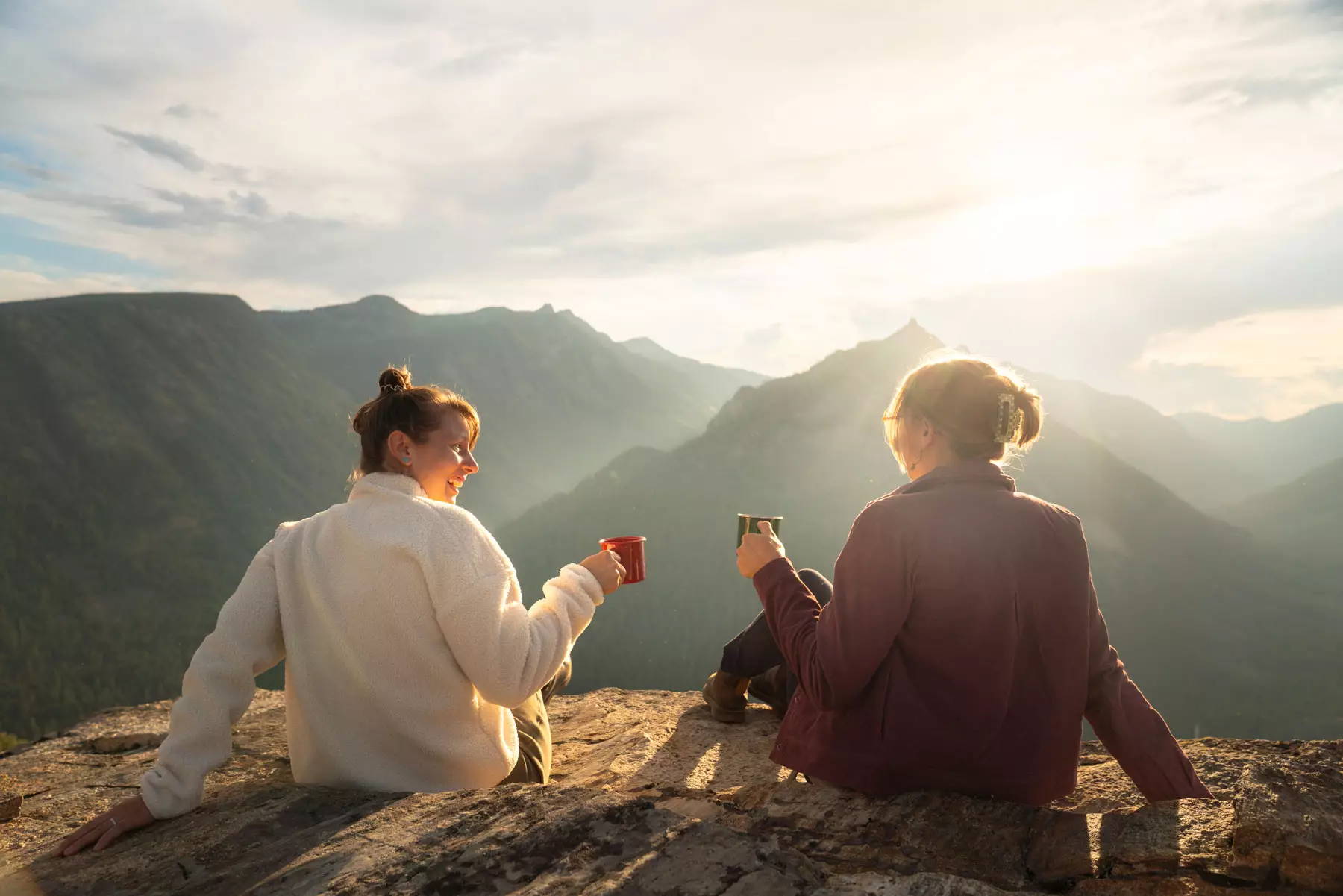 Two women sitting on cliff edge with coffee cups overlooking valley with sunrise.