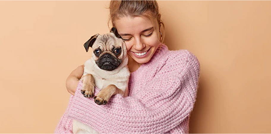 Smiling person in a sweater holding a cute pug against a beige background.