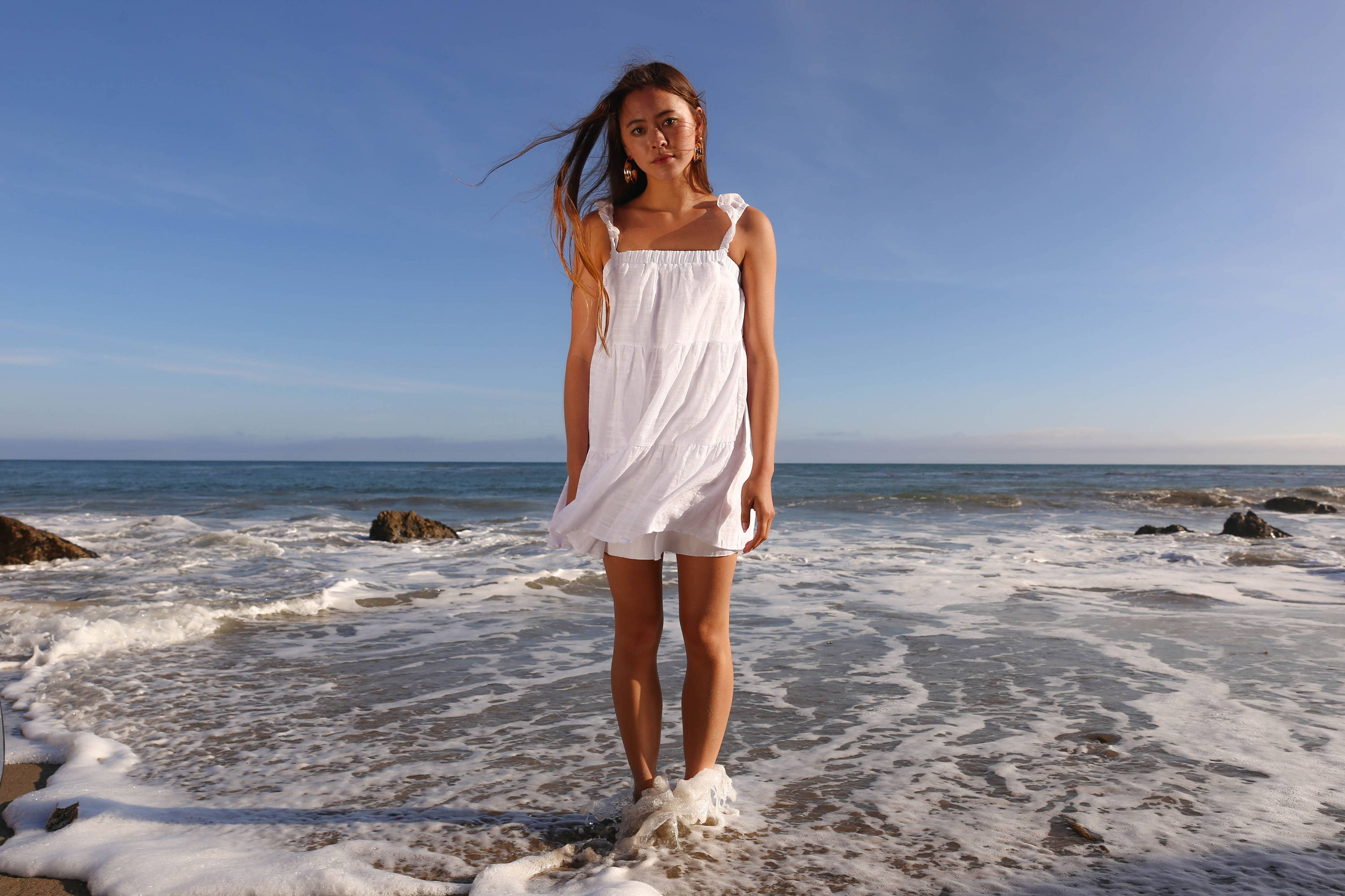 Trixxi Las Fotos Class C.E.O project photoshoot, girl standing in beach ocean water with white shoulder tie tiered short dress on.