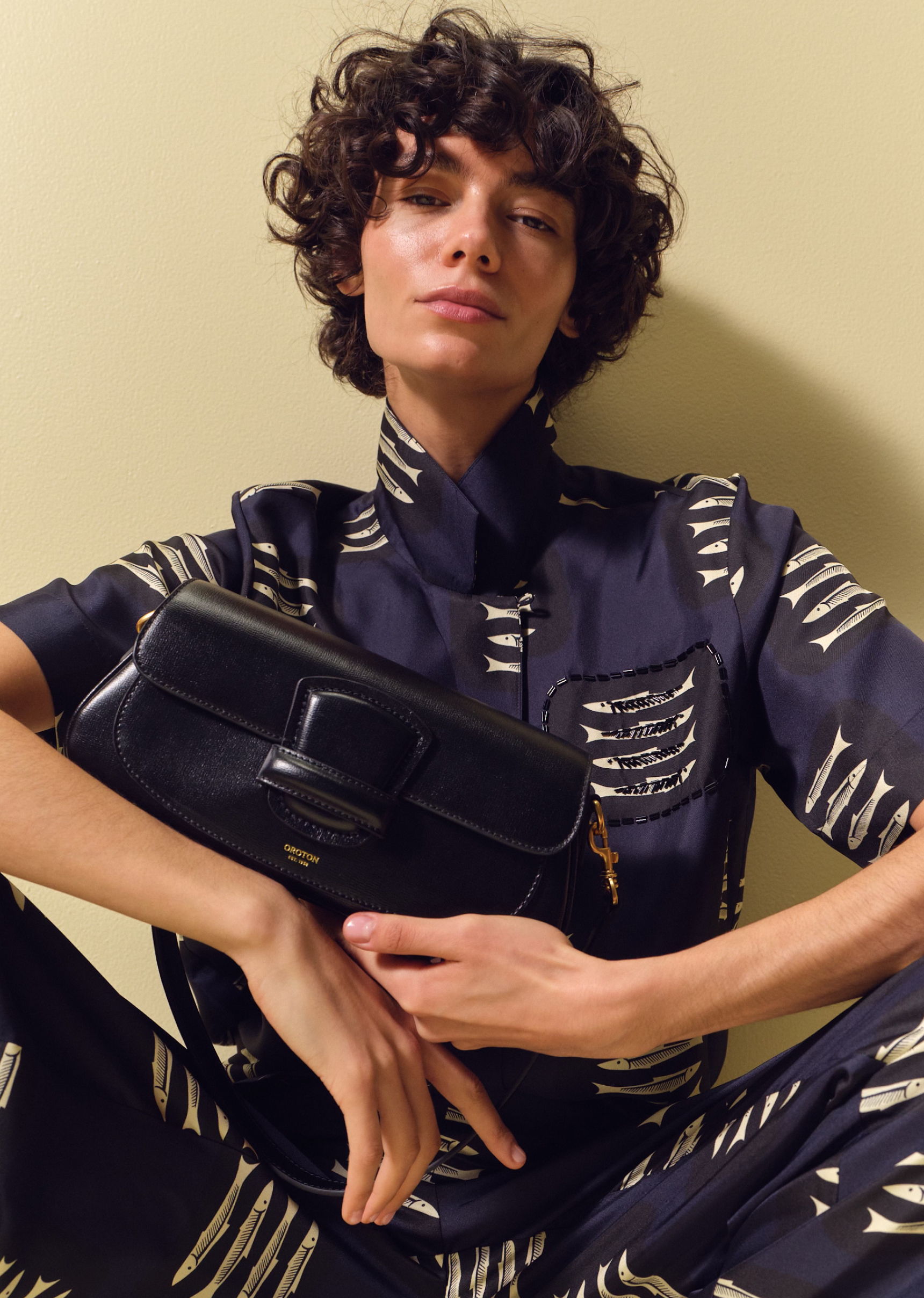 Person in patterned outfit holding a black handbag against a plain background.