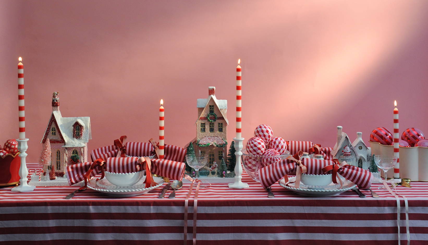 A wide image of a pink and red chirstmas laid table. With candles, crackers and christmas houses.