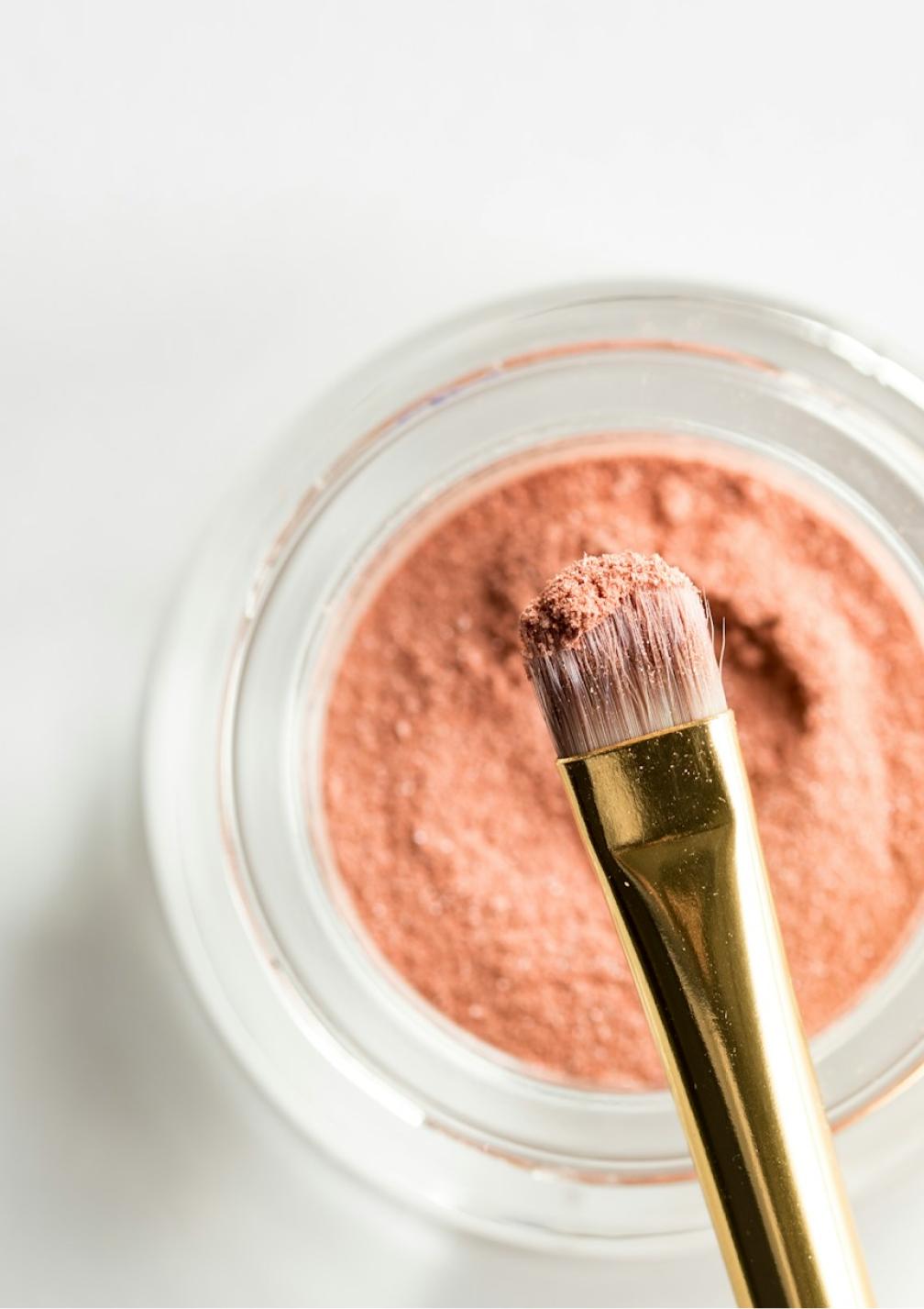 A glass pot containing makeup powder, with a gold-clad brush hovering over it, viewed top down.