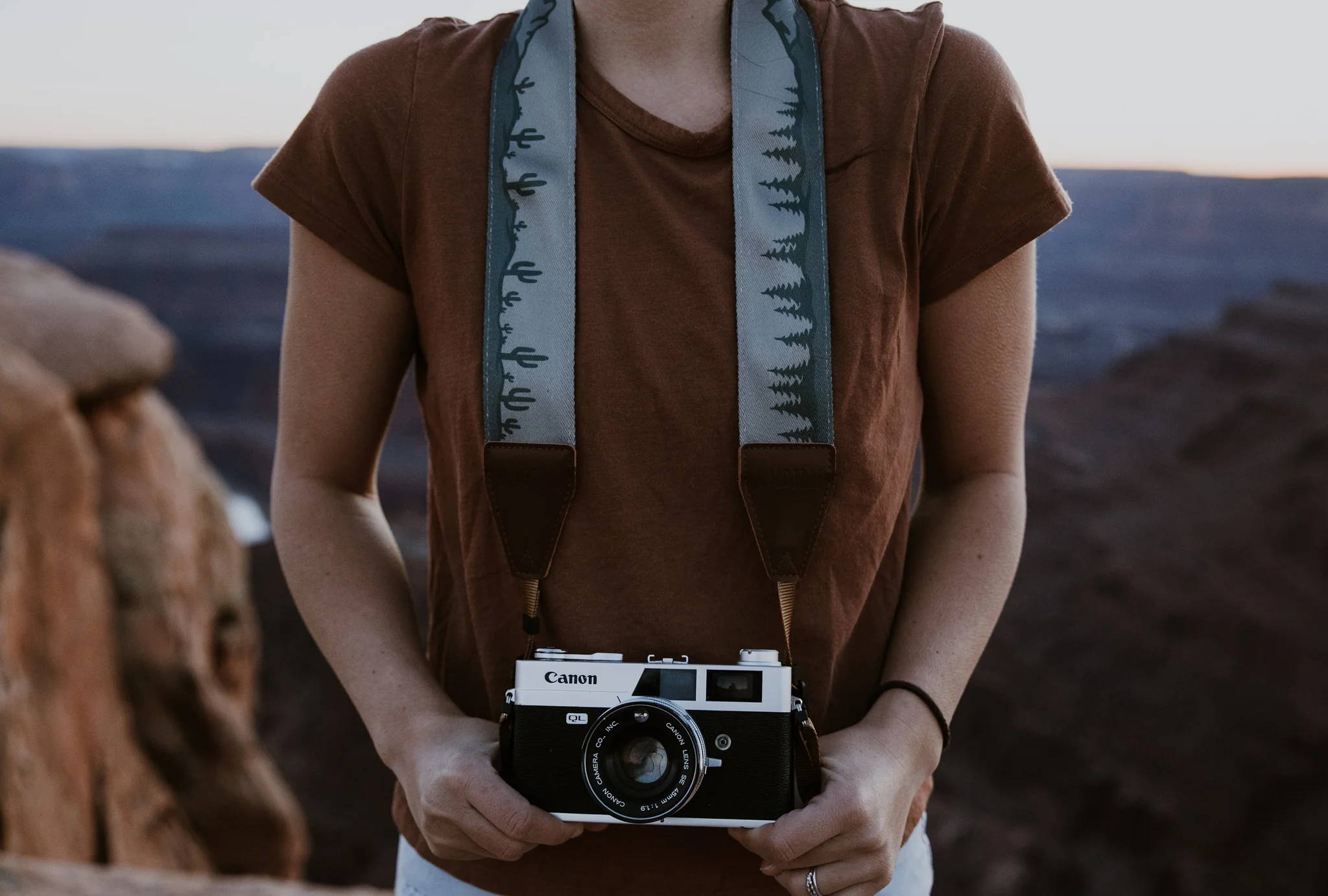 Woman holding a camera and wearing a camera neck strap in front of nature background.