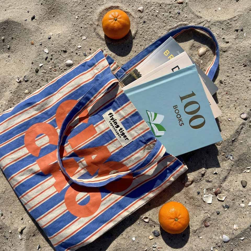 A striped tote bag with books and oranges on sandy beach.
